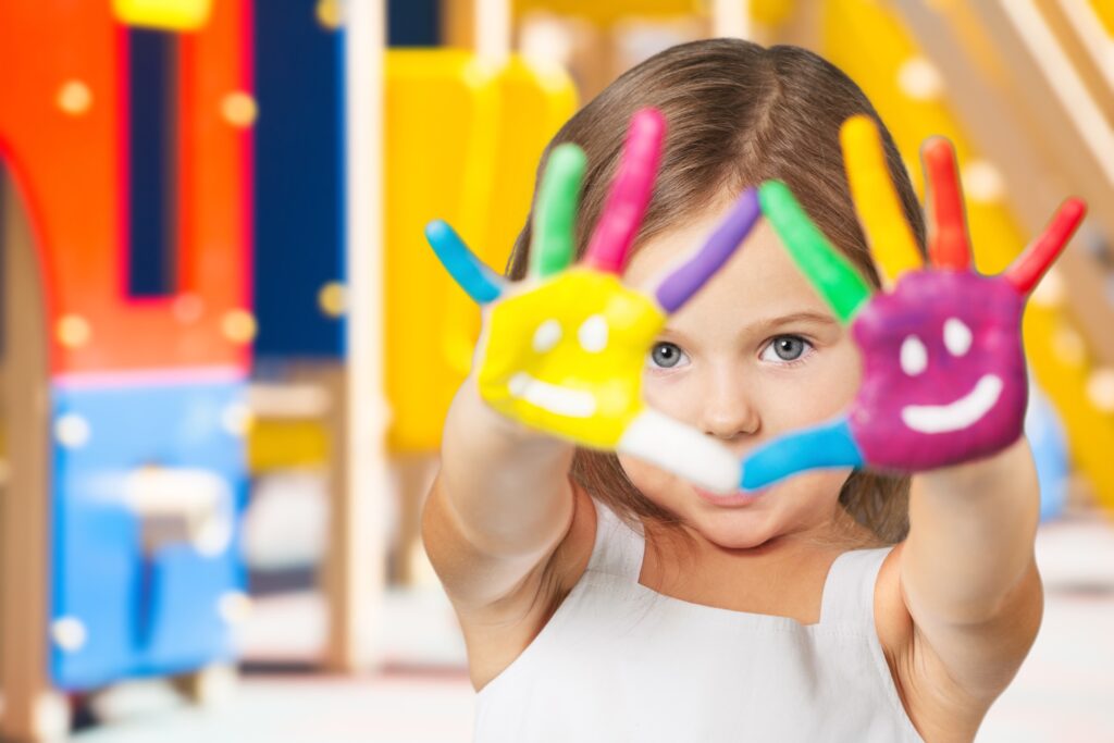 Child. Ein kleines Mädchen blickt durch ihre nach vorne gestreckten Hände, die bunt mit Fingermalfarbe und aufgemalten Smiley-Gesichtern verziert sind.