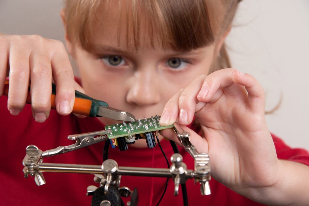 Childhobby Ein junges Mädchen arbeitet hochkonzentriert mit einem Seitenschneider an einer kleinen, in einer Halterung eingespannten Leiterplatte.