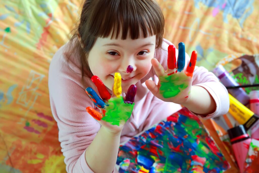 Cutelittlegirlwithpaintedhands Ein lächelndes junges Mädchen mit Down-Syndrom zeigt ihre bunt mit Fingerfarben bemallten Hände in die Kamera.