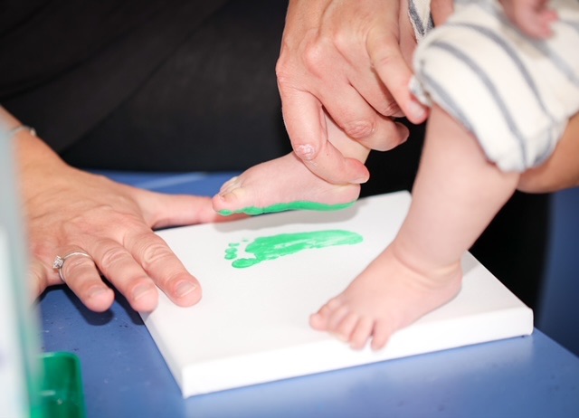 Eine erwachsene Person führt vorsichtig den mit grüner Farbe bemälten Fuß eines Babys auf eine weiße Leinwand, um einen Fußabdruck zu machen.