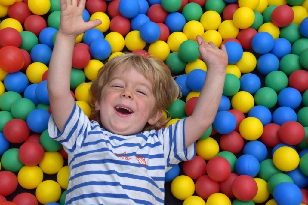 KleinerJungespieltimBällebad Ein kleiner blonder Junge liegt lachend auf dem Rücken in einem bunten Bällebad und streckt die Arme nach oben.