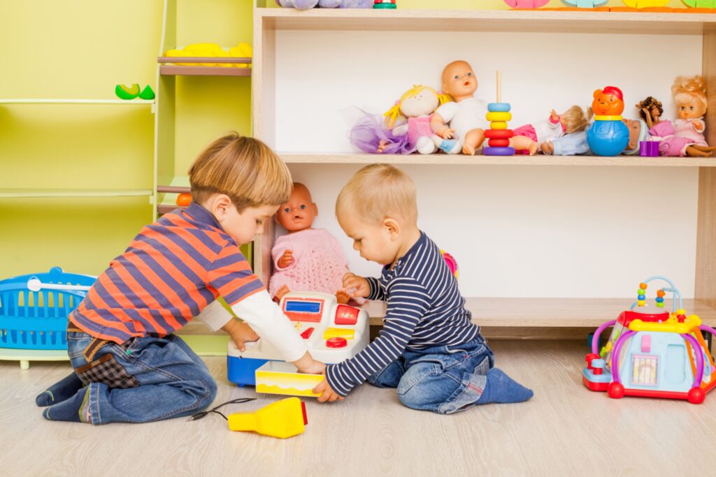 Littlecashierandbuyer Zwei kleine Jungen sitzen auf dem Boden eines Kinderzimmers und spielen gemeinsam Kaufladen mit einer Spielzeugkasse.