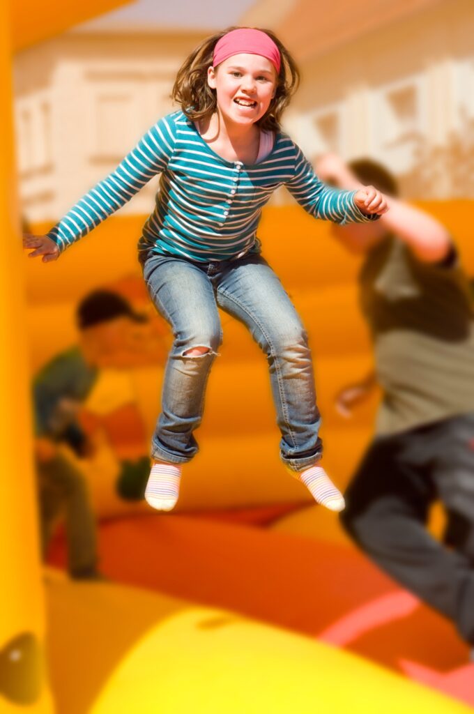 MädchenspringtaufderHüpfburg Ein junges Mädchen mit pinkem Stirnband springt mit einem fröhlichen Lachen hoch in der Luft auf einer gelb-orangefarbenen Hüpfburg.