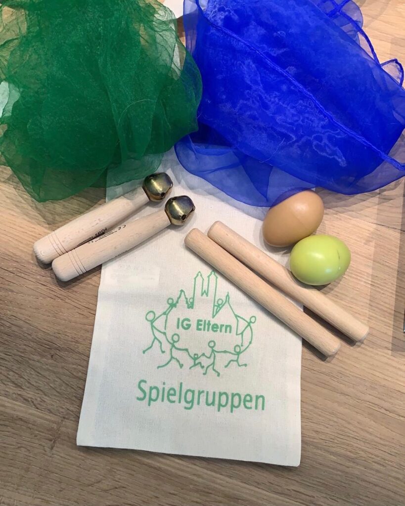 Musiksäckchen Ein kleines Stoffbeutelchen mit der Aufschrift „IG Eltern Spielgruppen“ liegt umgeben von Musik- und Spielutensilien wie Klanghölzern, Rassel-Eiern, Schellenstäben und bunten Tüchern auf einem Holzboden.