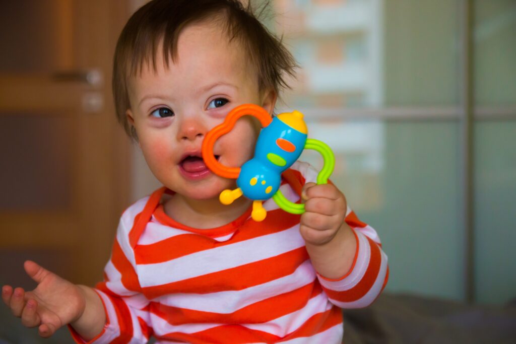 PortraitofcutebabyboywithDownsyndrome Ein glückliches Kleinkind mit Down-Syndrom sitzt in einem rot-weiß gestreiften Oberteil da und hält eine bunte Rassel in der Hand, an der es freudig knabbert.