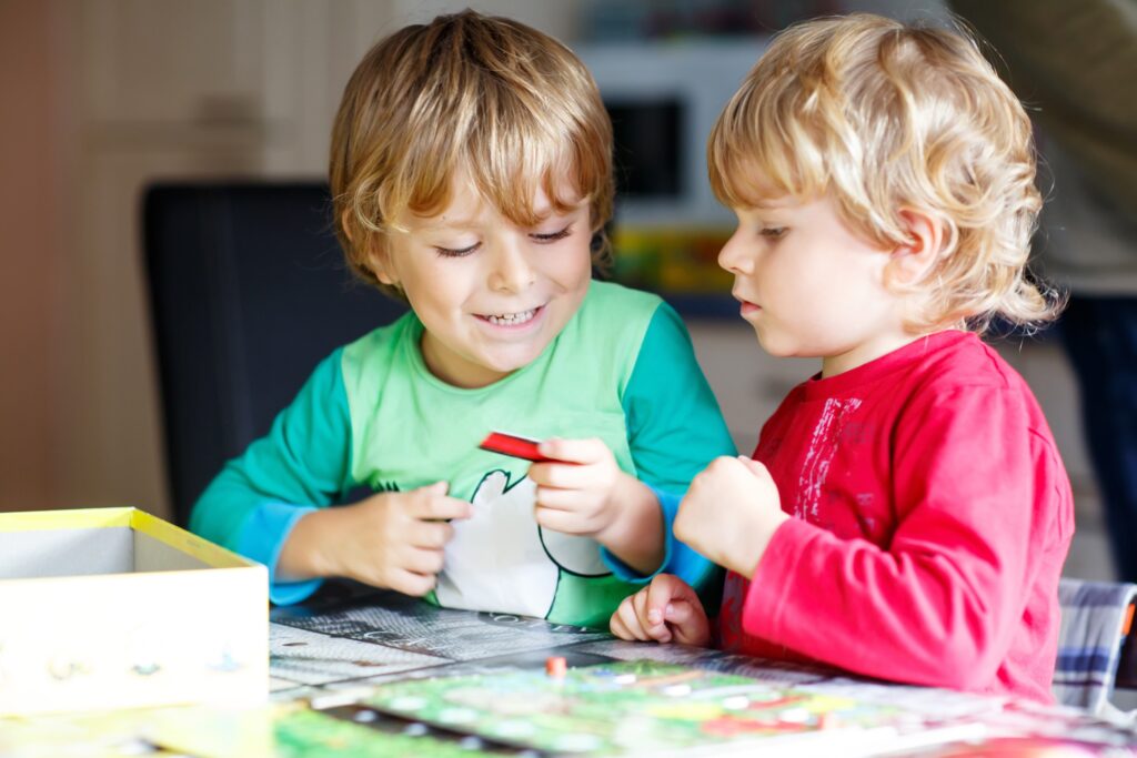 Twolittleblondkidboysplayingtogetherboardgameathome.Funnysiblingshavingfun. Zwei kleine, blonde Brüder sitzen am Tisch und spielen gut gelaunt ein Brettspiel, während der eine dem anderen stolz eine Spielkarte zeigt.
