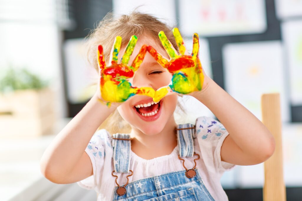 funnychildgirldrawslaughingshowshandsdirtywithpaint Ein kleines Mädchen lacht herzlich und formt mit ihren bunt bemälten Händen ein Herz vor ihren Augen.