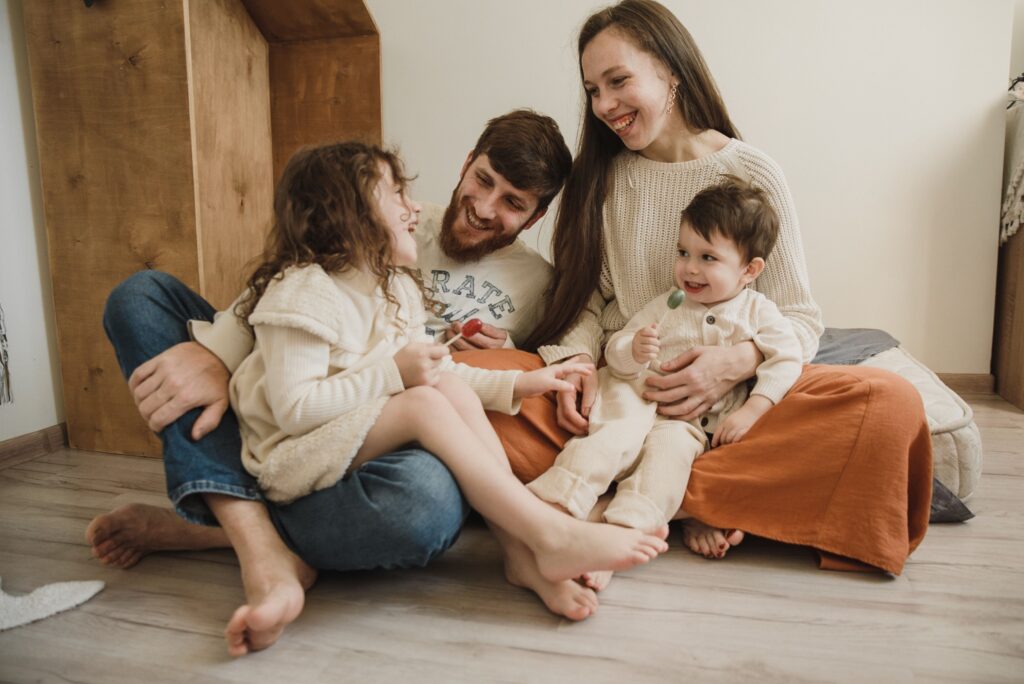 pexels-arina-krasnikova-5103918 Eine vierköpfige Familie sitzt barfuß und lachend zusammen auf dem Zimmerboden, während die beiden kleinen Kinder Lutscher in den Händen halten.