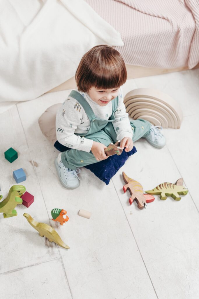 pexels-cottonbro-3661337 Ein kleines Kind mit braunem Topfschnitt sitzt auf dem Boden und spielt fröhlich mit verschiedenen Dinosaurier-Spielfiguren aus Holz.