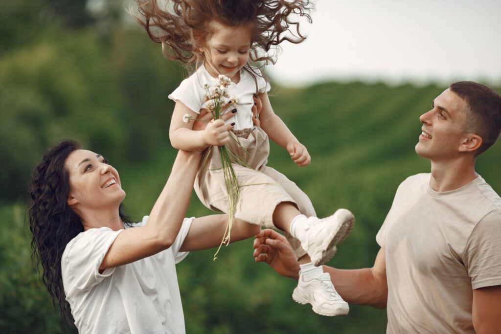 pexels-gustavo-fring-4971046 Eine glückliche Familie im Freien: Eine Mutter hebt ihre kleine Tochter lachend in die Luft, während der Vater daneben steht und beide lächelnd ansieht.