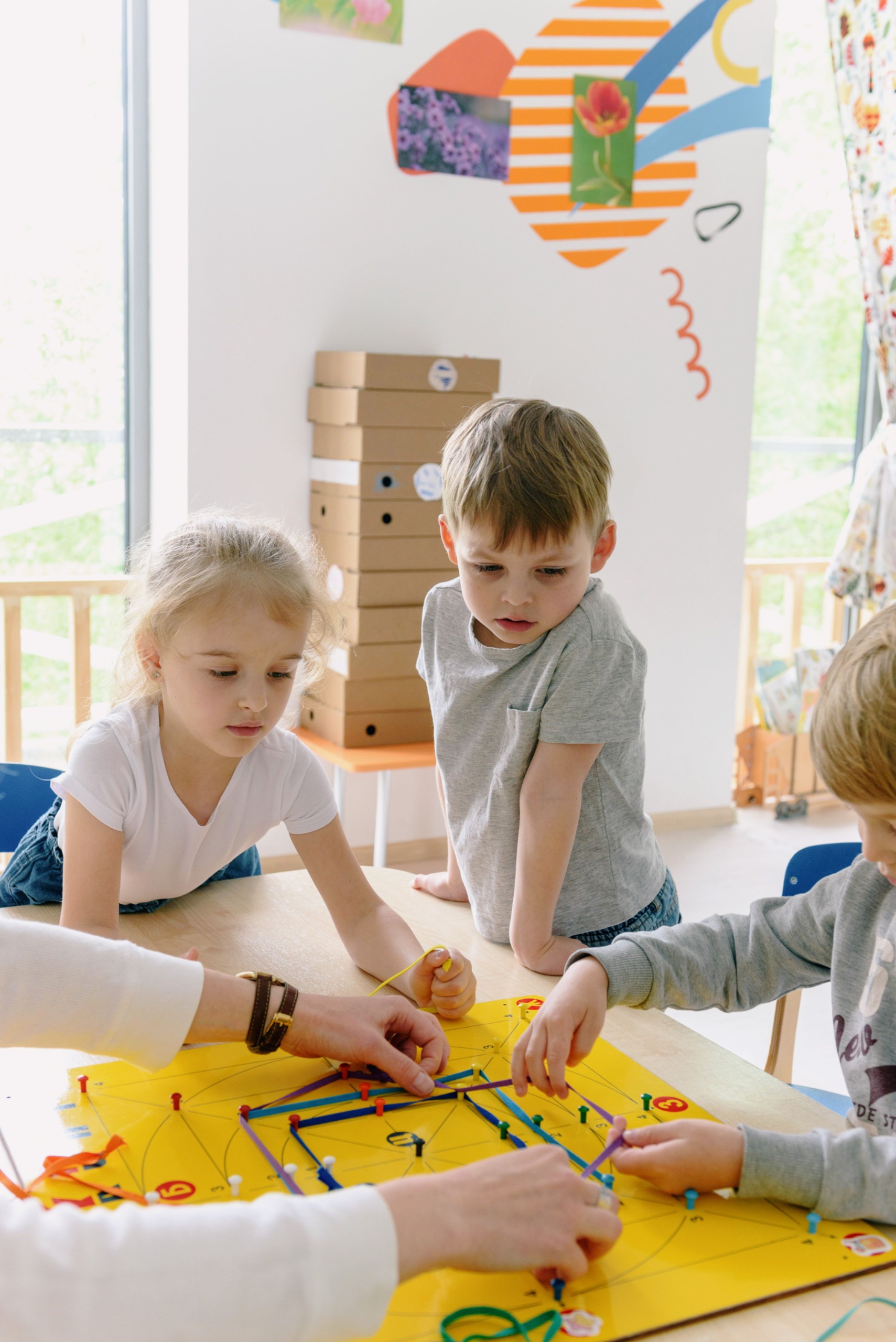 Drei Kinder sitzen und stehen konzentriert um einen Tisch und spannen gemeinsam mit einer erwachsenen Person bunte Bänder auf ein gelbes Lernspielbrett.