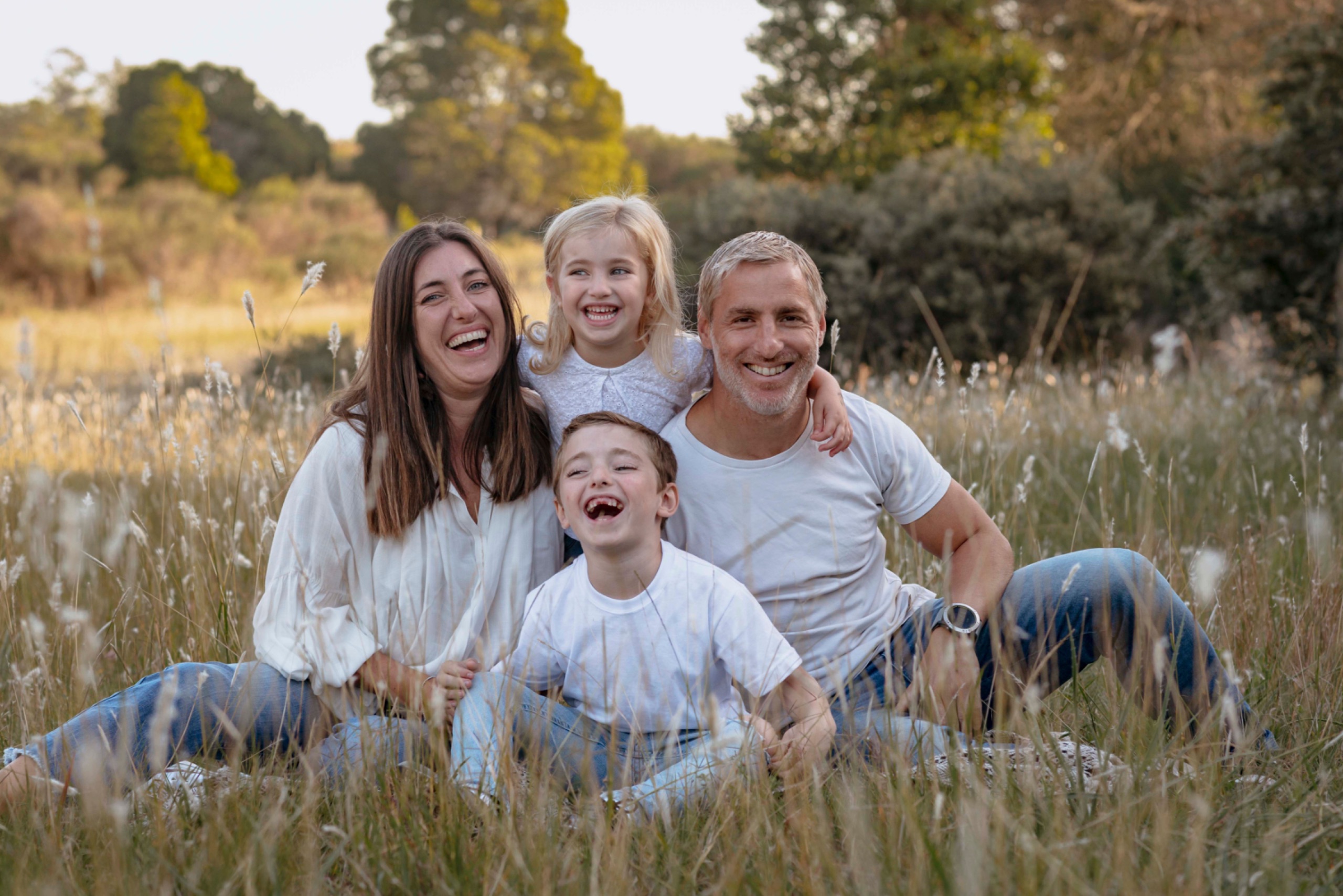 Eine vierköpfige Familie sitzt glücklich und herzlich lachend in einer hohen Graswiese im Freien.