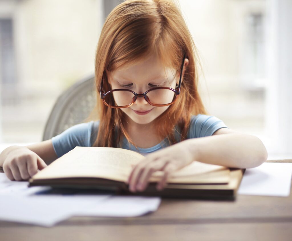 pexels-photo-3768122-3768122 A young girl with glasses intently reads a book indoors, embodying the essence of learning and discovery.