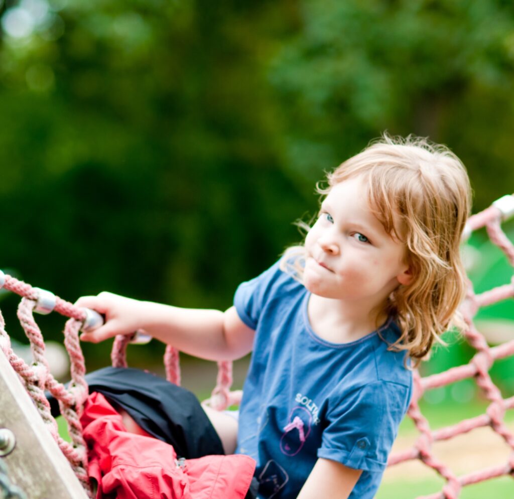 schaukeln Ein kleines Kind mit rötlich-blonden Haaren sitzt auf einer großen Netzschaukel oder einem Kletternetz auf einem Spielplatz und blickt mit konzentriertem Gesichtsausdruck zur Seite.